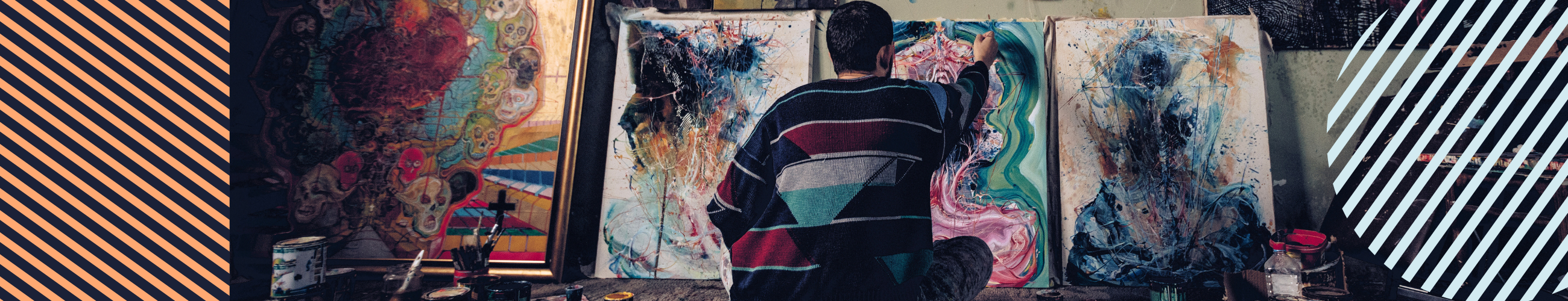 Masculine artist painting cross legged on the floor with paintings against the wall in front of them