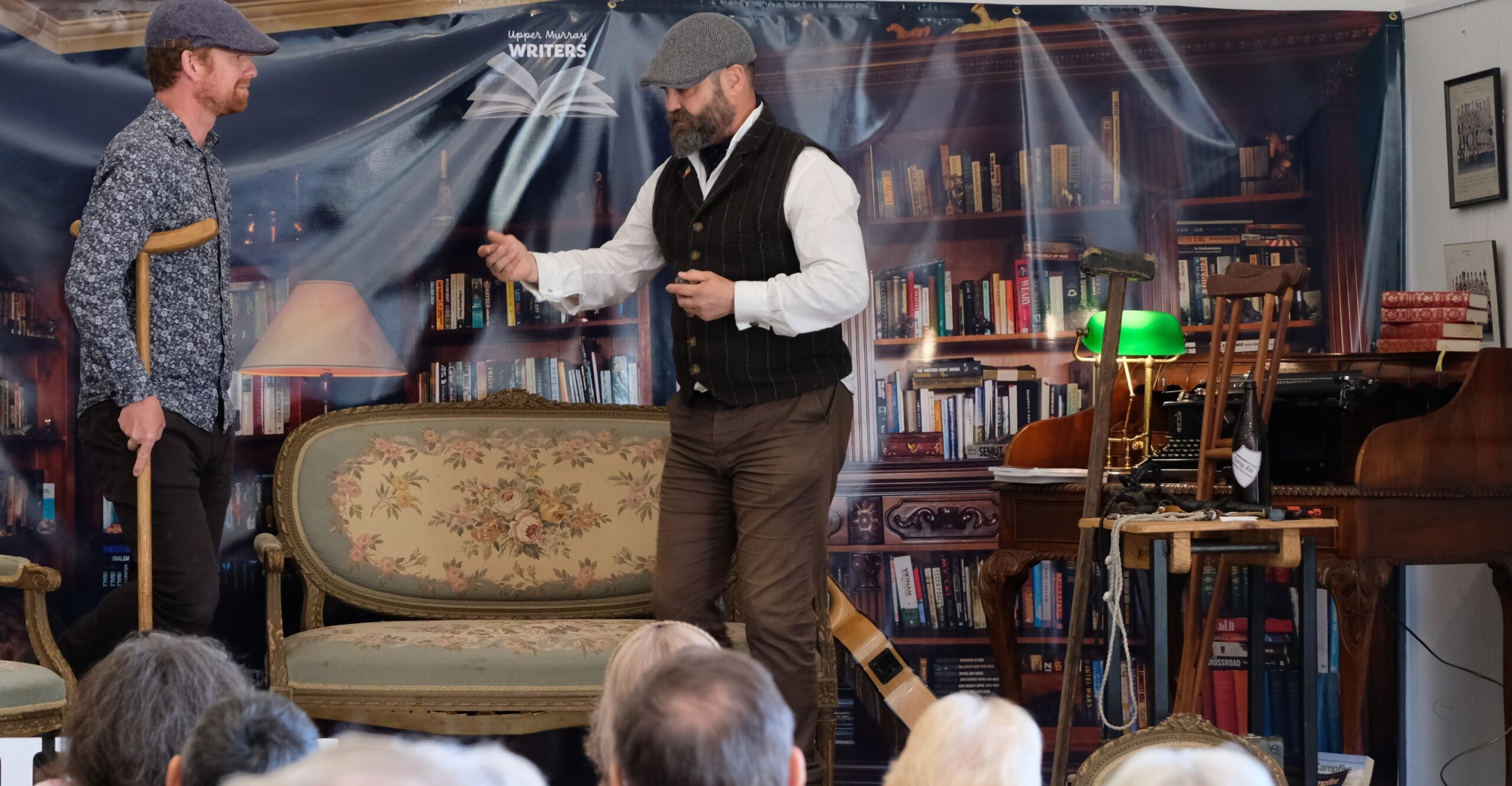 Two men standing in front of a bookcase next to a writing desk talking on a microphone.