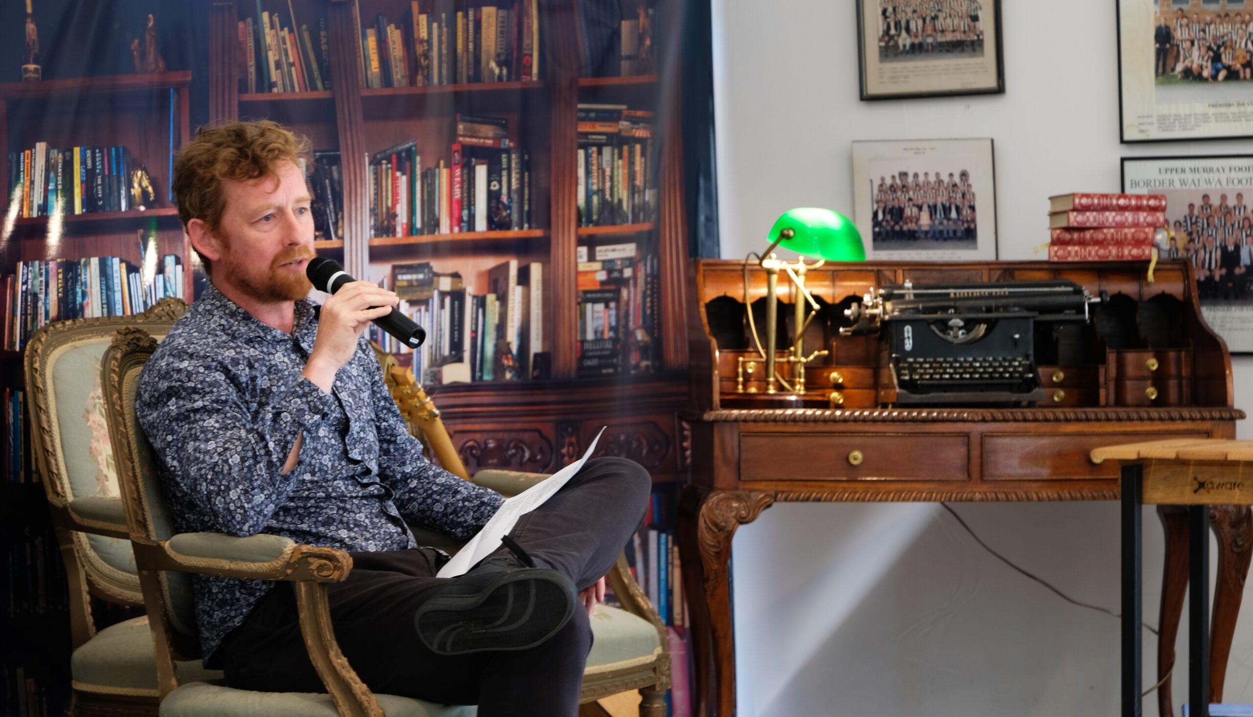 Man sitting front of a bookcase next to a writing desk talking on a microphone.