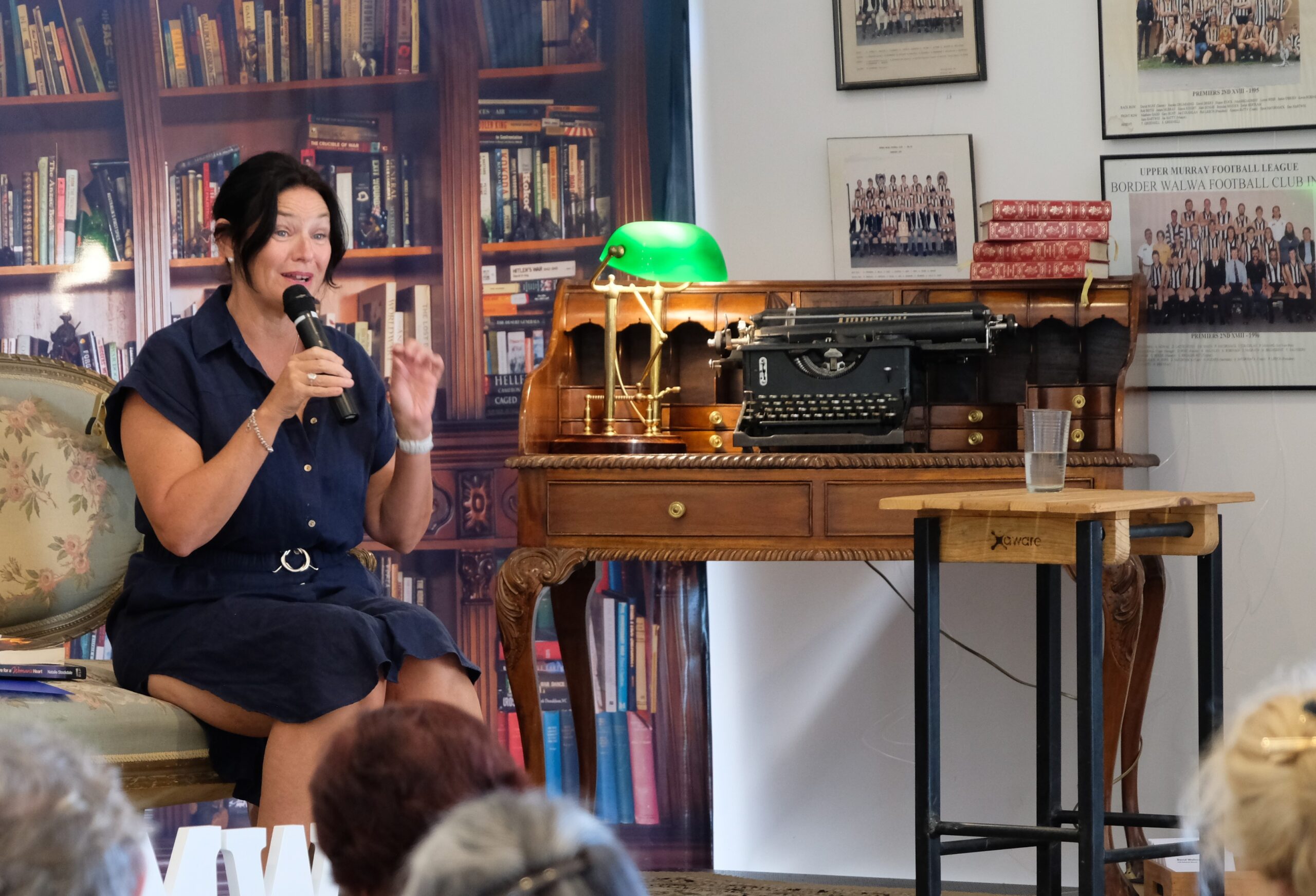 Woman sitting front of a bookcase next to a writing desk talking on a microphone.