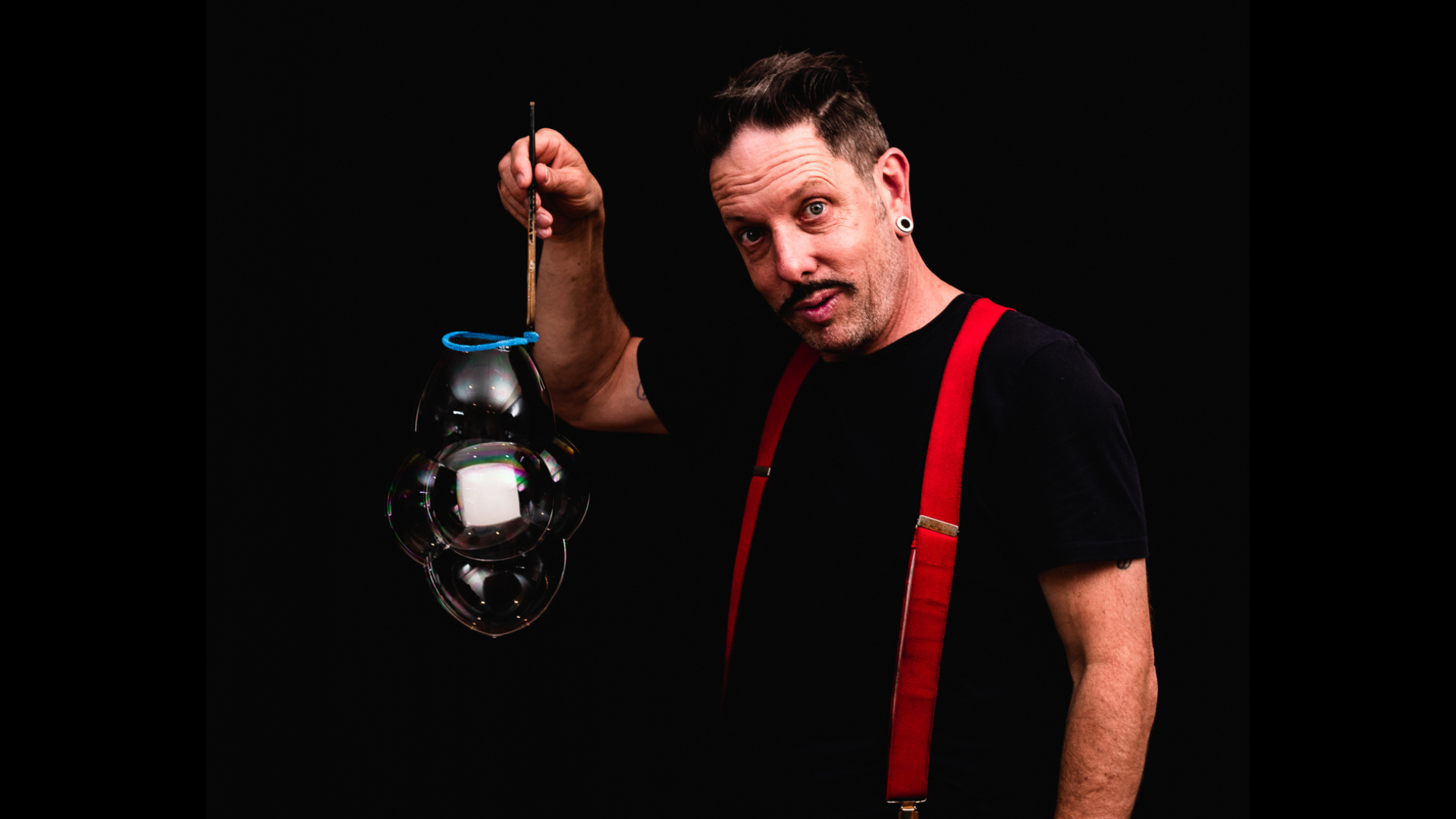 Man in black shirt and red suspenders holding a large bubble.